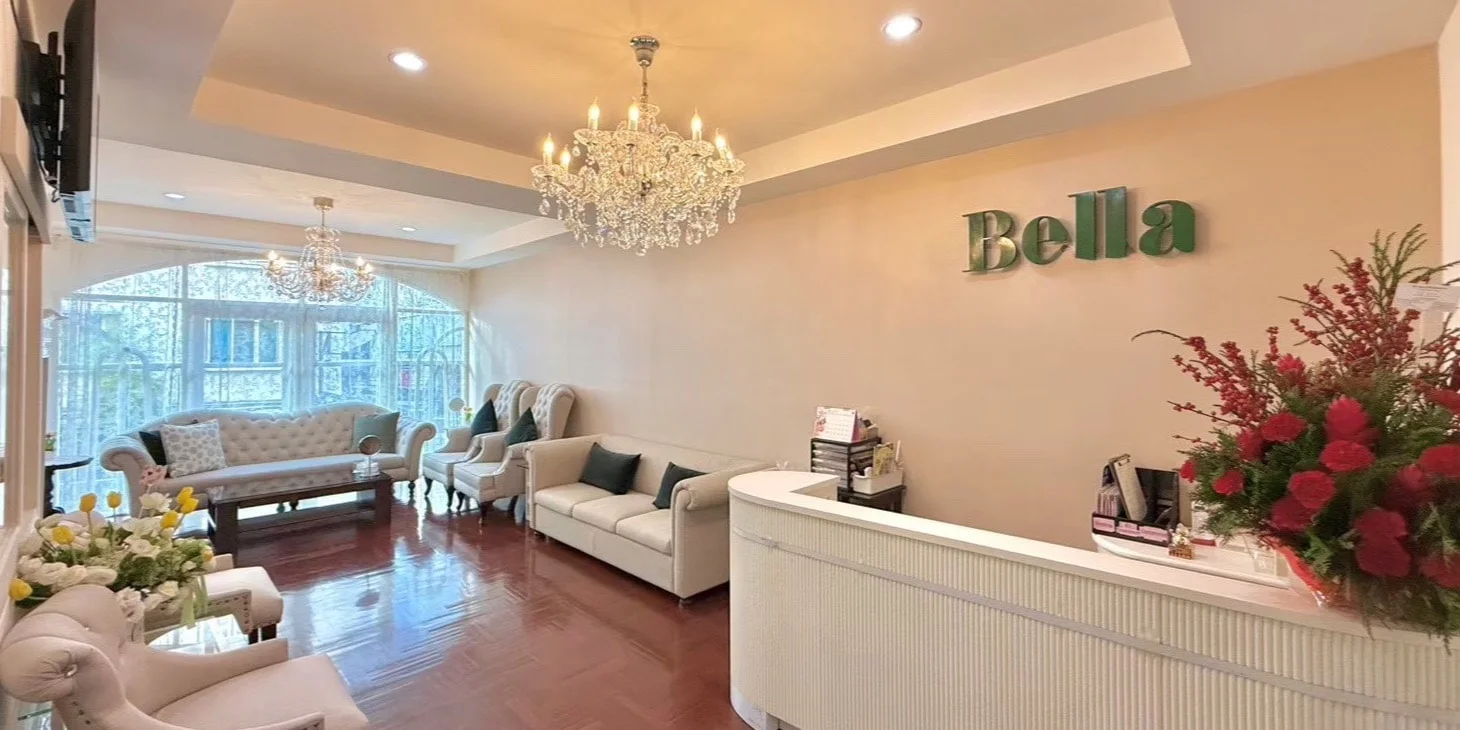ห้องรับรองของ Bella Clinic เคาน์เตอร์สีงาช้าง ป้าย Bella สีเขียวบนผนัง โคมระย้าคริสตัล และโซฟาสีครีม